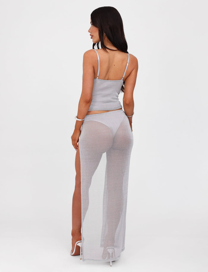 A woman with long dark hair stands against a plain white background, dressed in the WYNNE SKIRT - GREY : SILVER from TIGERMIST. She pairs the light gray, sheer, asymmetrical maxi skirt featuring a low-rise waist with a matching crop top and white high-heeled sandals. She poses with her back to the camera, glancing over her shoulder.