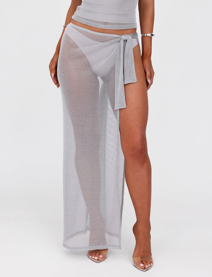 A person wearing the TIGERMIST WYNNE SKIRT in grey/silver, a sheer wrap skirt that ties at the low-rise waist. The skirt is paired with transparent high-heeled shoes, a fitted top that partially shows, and minimal jewelry, including a bracelet. The background is plain white.
