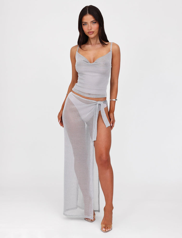 A woman with long, dark hair wears a sheer two-piece outfit from TIGERMIST. The top is a spaghetti-strap camisole, and the bottom is the WYNNE SKIRT - GREY : SILVER, a metallic knit wrap skirt with a high slit that reveals a bikini underneath. Featuring a low-rise waist that accentuates her figure, she confidently poses against a plain white background.