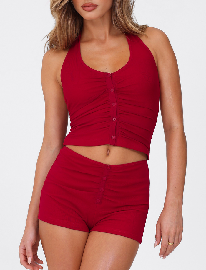A woman models the TIGERMIST ZAFIRA SHORT - RED, a body-con bright red mini short, paired with a matching sleeveless ruched crop top, confidently posing against a plain white background.