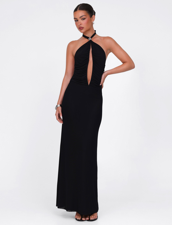 A woman stands against a white background wearing the TIGERMIST ZARA MAXI DRESS in black—a sleeveless halter style with front keyhole cutout and high-stretch, figure-flattering fabric. She accessorizes with earrings, a bracelet, and hair styled back.