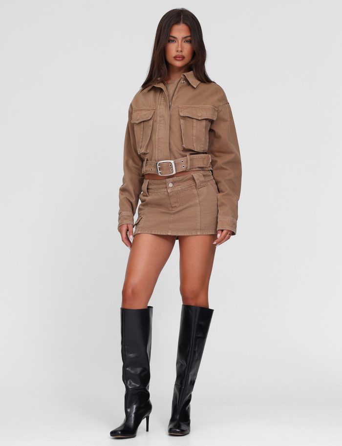 A woman with long brown hair and a neutral expression stands against a plain background, wearing the TIGERMIST ZELIE MINI SKORT in washed tan with a matching denim jacket and knee-high black heeled boots.