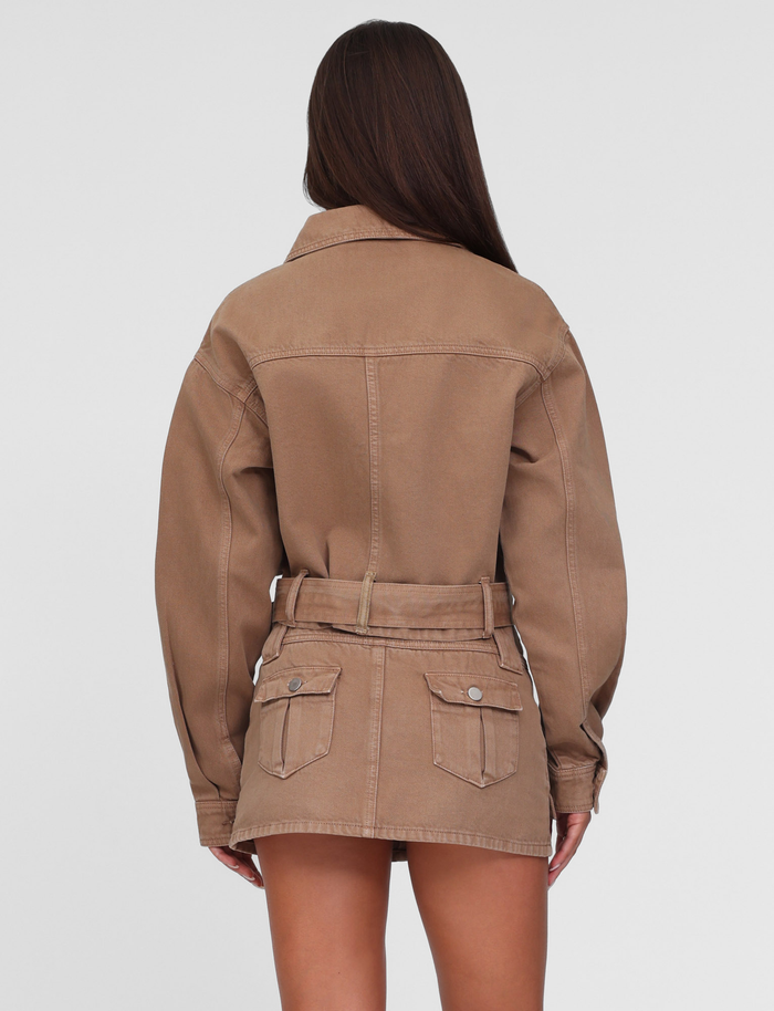 A woman with long brown hair faces away, wearing the TIGERMIST ZELIE MINI SKORT in Neutral Sandblast Washed Tan with functional pockets and a matching jacket, standing against a plain white background.