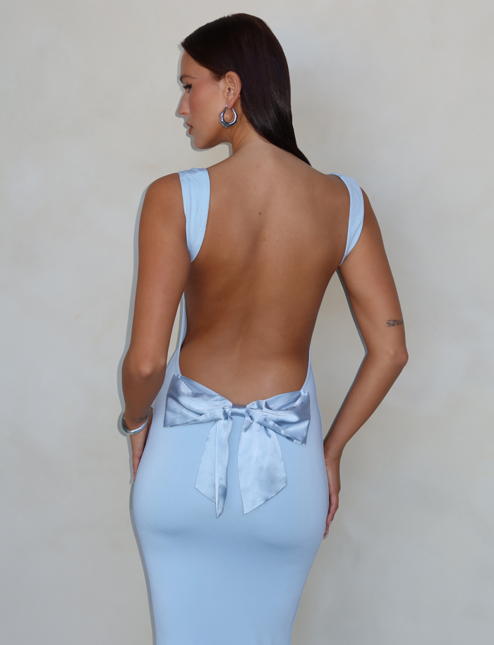 A woman faces away from the camera in the TIGERMIST ZITA DRESS - BLUE: BABY BLUE, showcasing its elegant open back with a satin bow and flattering bodycon silhouette.