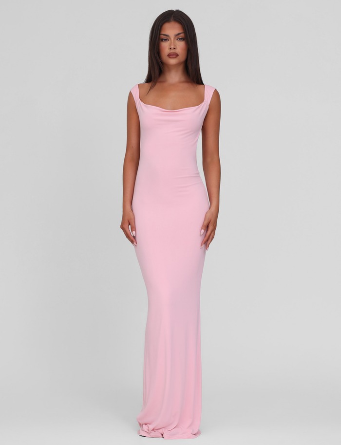 A woman with long straight brown hair stands against a light gray background, wearing the TIGERMIST ZITA DRESS in Baby Pink. This long, sleeveless bodycon dress features a draped neckline. She has a neutral expression and arms relaxed at her sides.