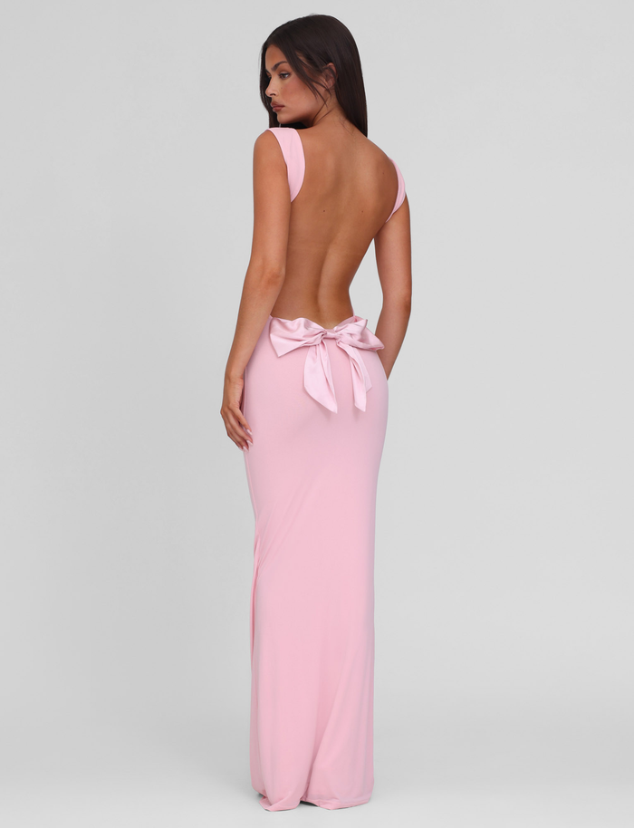 A woman models the TIGERMIST ZITA DRESS - PINK : BABY PINK, a pale pink maxi gown with wide straps, backless design, and a large bow at the lower back, standing against a plain light background and looking over her shoulder.